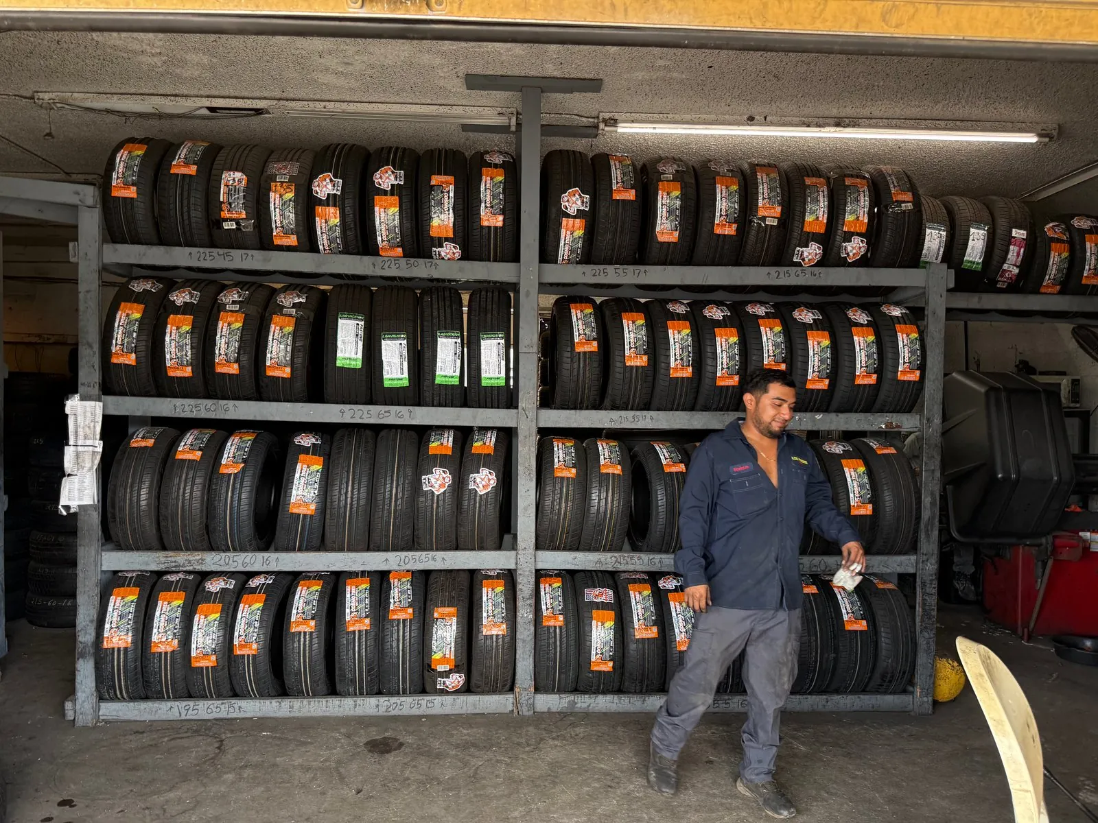 Estantes de llantas nuevas con etiquetas del fabricante en la banda de rodadura, ordenadas por medida, y un técnico de Los Paisas en el taller